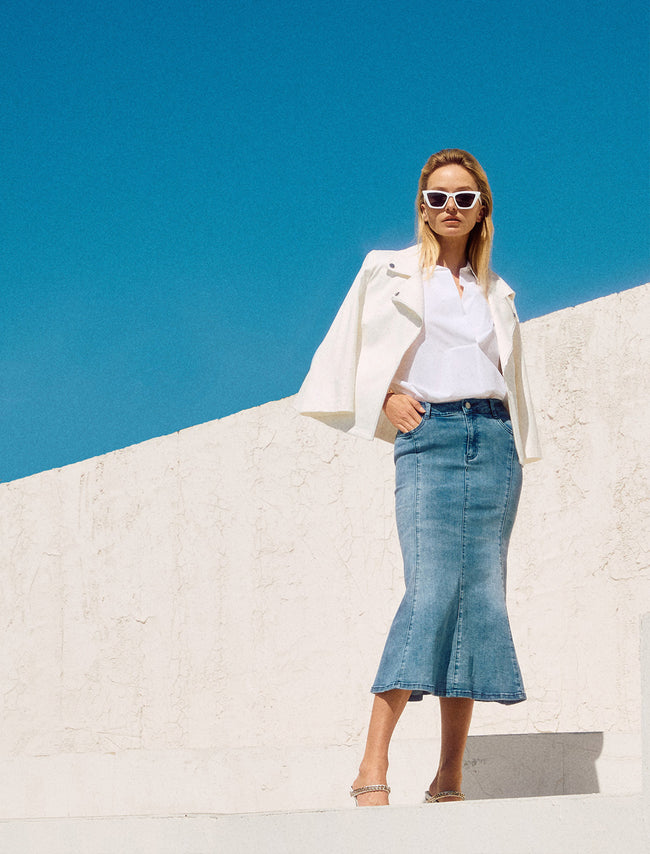 Joseph Ribkoff Banner mit eleganter Frau in weißem Blazer, weißer Bluse und blauem Jeansrock, kombiniert mit Sonnenbrille und Sandaletten, aufgenommen vor moderner Architektur mit weißer Steinwand und strahlend blauem Himmel.