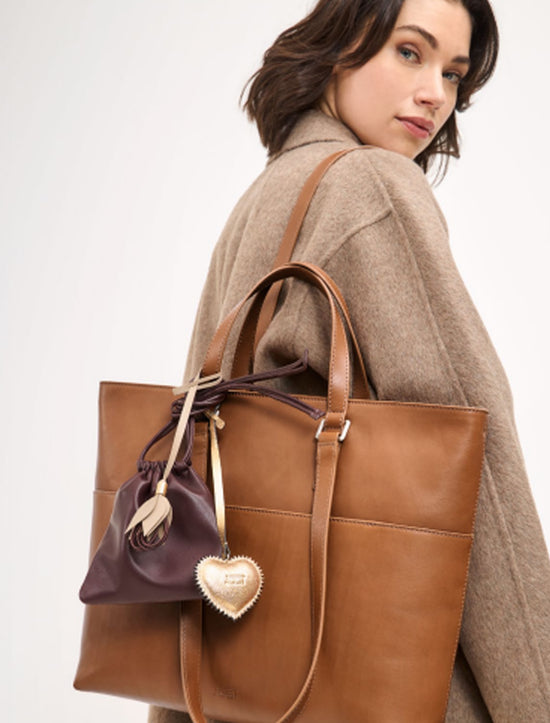 Banner für Jost Taschen Wien Herbst Winter 2025. Eine Frau in beigefarbenem Wollmantel trägt eine große, eckige Schultertasche aus glattem cognacbraunem Leder. An der Tasche hängt ein kleines dunkelviolettes Beutelchen mit goldenem Herzanhänger als charmantes Detail. Der Look kombiniert dezente Farben mit hochwertigem Material und unterstreicht die elegante, moderne Ästhetik der Marke Jost.