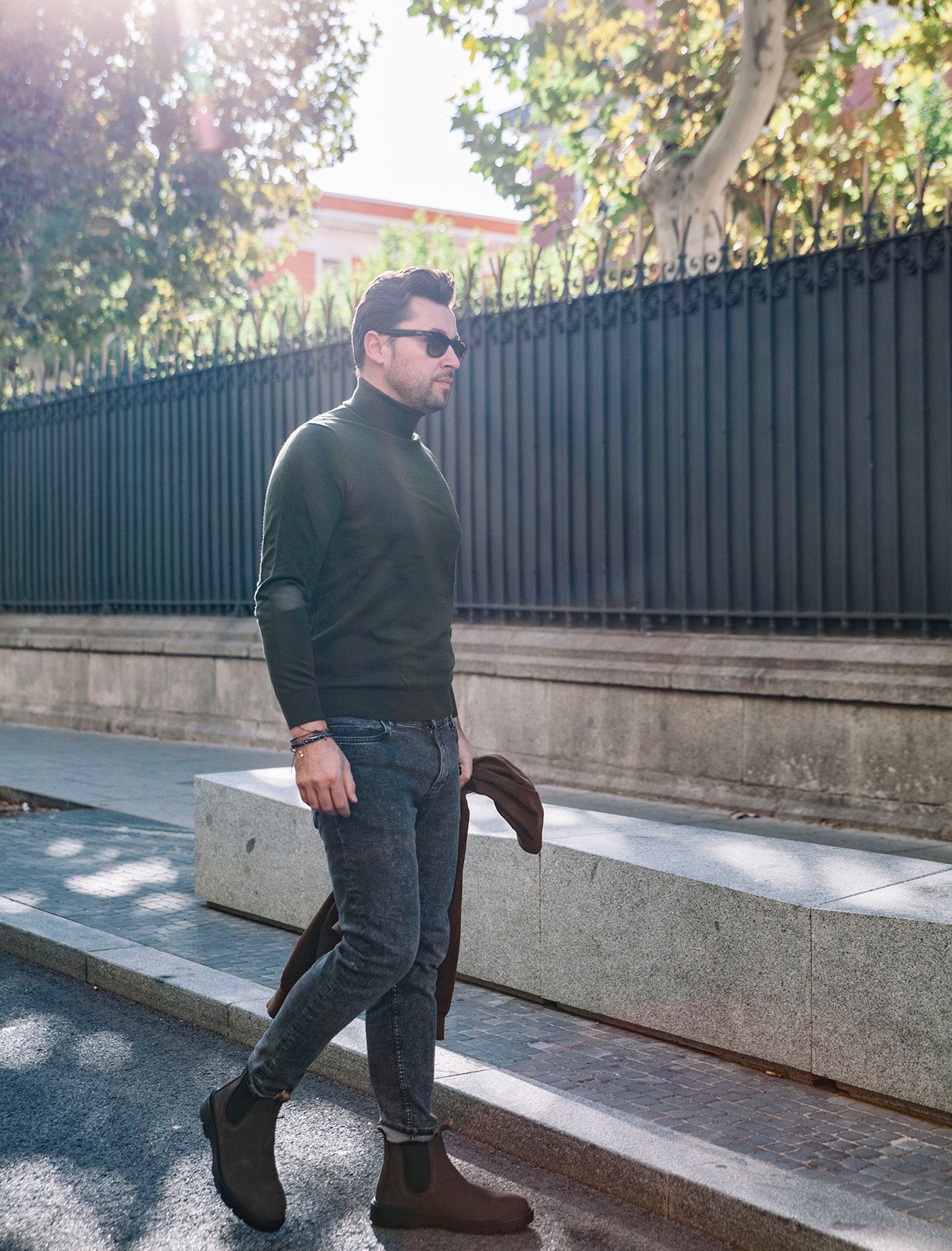 Natural World Banner mit stilvollem Herren-Outfit in der Stadt. Ein Mann trägt braune nachhaltige Boots aus Leder im Chelsea-Stil mit elastischen Einsätzen, kombiniert mit schmaler grauer Jeans, dunkelgrünem Rollkragenpullover und Sonnenbrille. In der Hand hält er eine Jacke, während er auf einem Gehweg mit Steinmauern und schwarzem Metallzaun entlanggeht. Sonnenlicht und Bäume im Hintergrund erzeugen eine moderne Streetstyle-Atmosphäre.