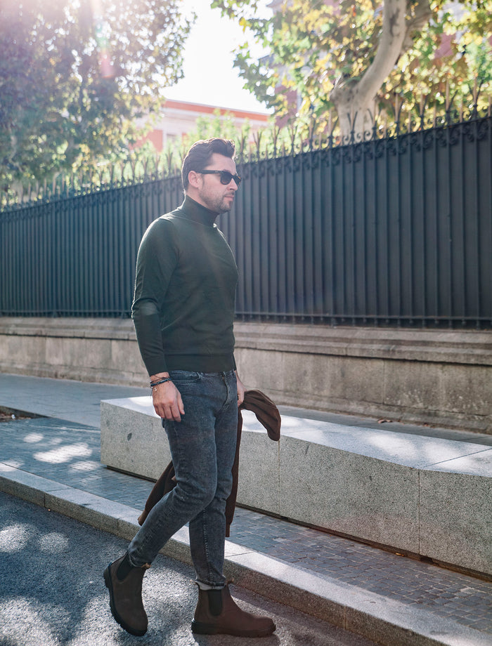 Natural World Banner mit stilvollem Herren-Outfit in der Stadt. Ein Mann trägt braune nachhaltige Boots aus Leder im Chelsea-Stil mit elastischen Einsätzen, kombiniert mit schmaler grauer Jeans, dunkelgrünem Rollkragenpullover und Sonnenbrille. In der Hand hält er eine Jacke, während er auf einem Gehweg mit Steinmauern und schwarzem Metallzaun entlanggeht. Sonnenlicht und Bäume im Hintergrund erzeugen eine moderne Streetstyle-Atmosphäre.