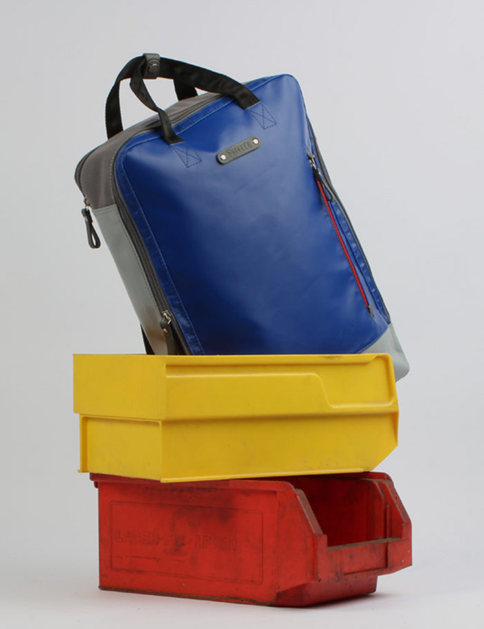 7Clouds Banner mit nachhaltigem Rucksack in Blau und Grau, gefertigt aus recycelten Materialien im Upcycling-Design. Der Rucksack mit schwarzen Trageriemen und seitlichem Reißverschlussfach ist auf zwei bunten Kunststoffboxen in Gelb und Rot drapiert. Minimalistischer Hintergrund in Weiß betont die Farben und unterstreicht den urbanen, nachhaltigen Charakter der Marke.