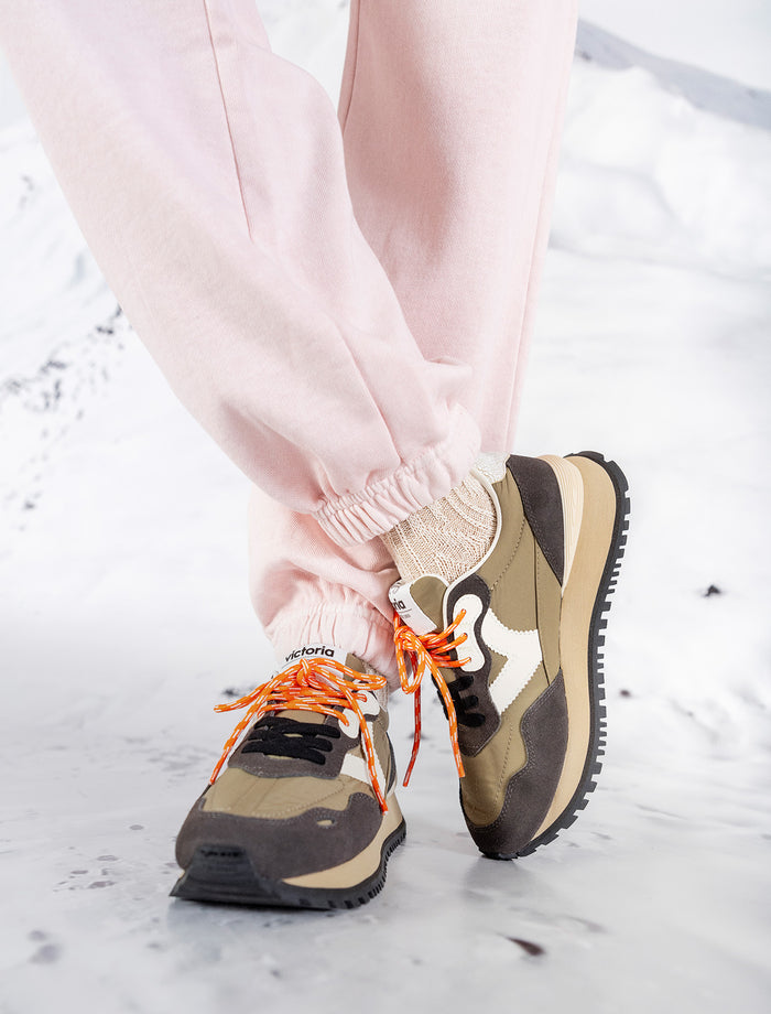 Banner mit Victoria Sneakern aus der Herbst Winter Kollektion 2025. Detailaufnahme von khakifarbenen Sneakers mit braunen und schwarzen Akzenten, weißem Markenlogo an der Seite und auffälligen orangefarbenen Schnürsenkeln. Kombiniert mit rosafarbenen Jogginghosen und beigen Socken, fotografiert auf weißem Untergrund mit winterlicher Anmutung. Der Look vereint sportlichen Komfort mit modernem Streetstyle.